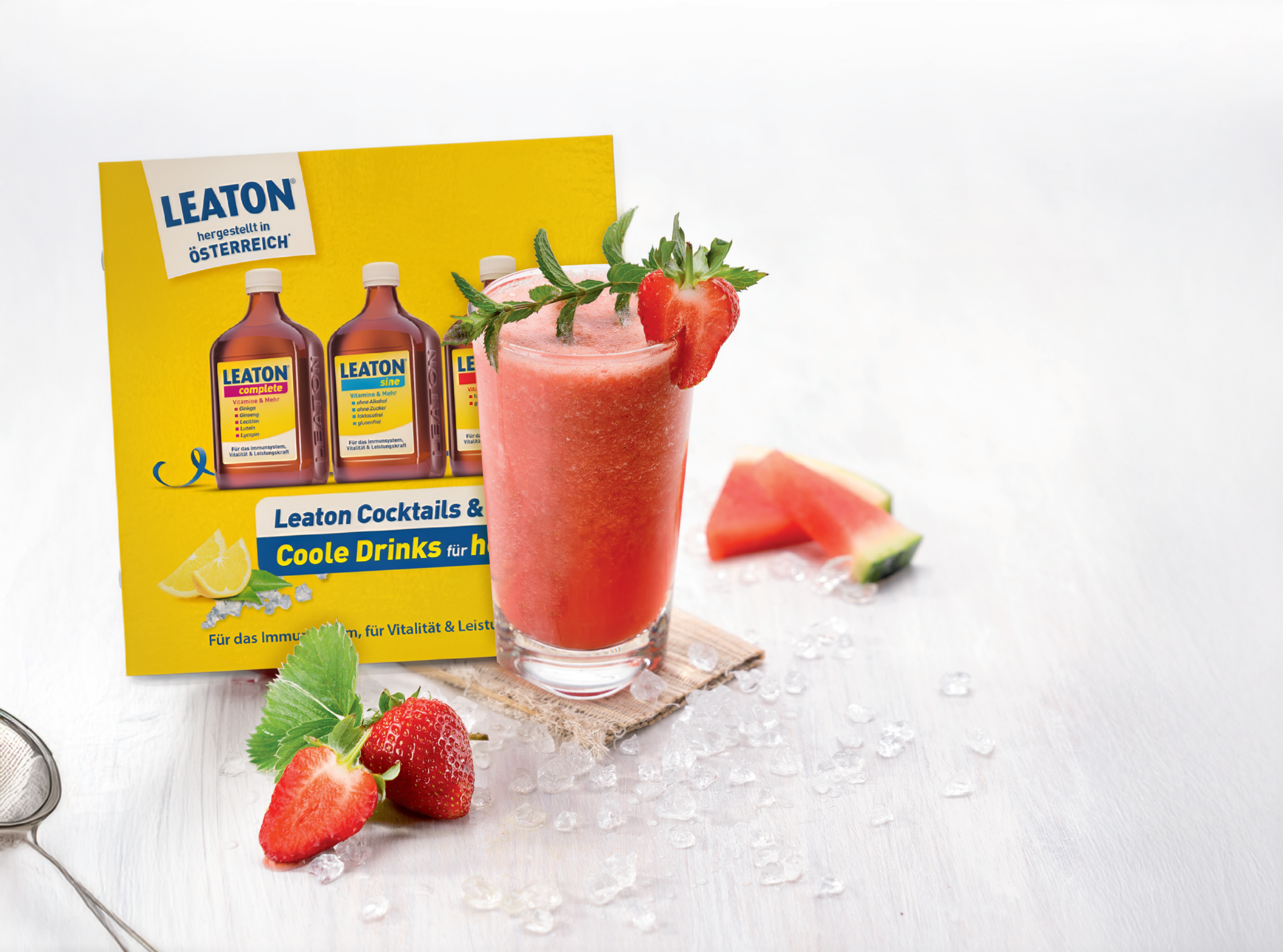 Ein rotes Getränk mit Minze und Erdbeere in einem Glas vor einer Leaton-Packung, umgeben von Eiswürfeln, Erdbeeren und Melonenscheiben.