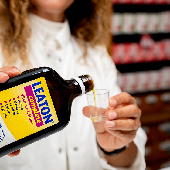 Eine Person in weißem Kittel gießt ein gelbes Vitaminpräparat aus einer beschrifteten Flasche in einen kleinen Messbecher.