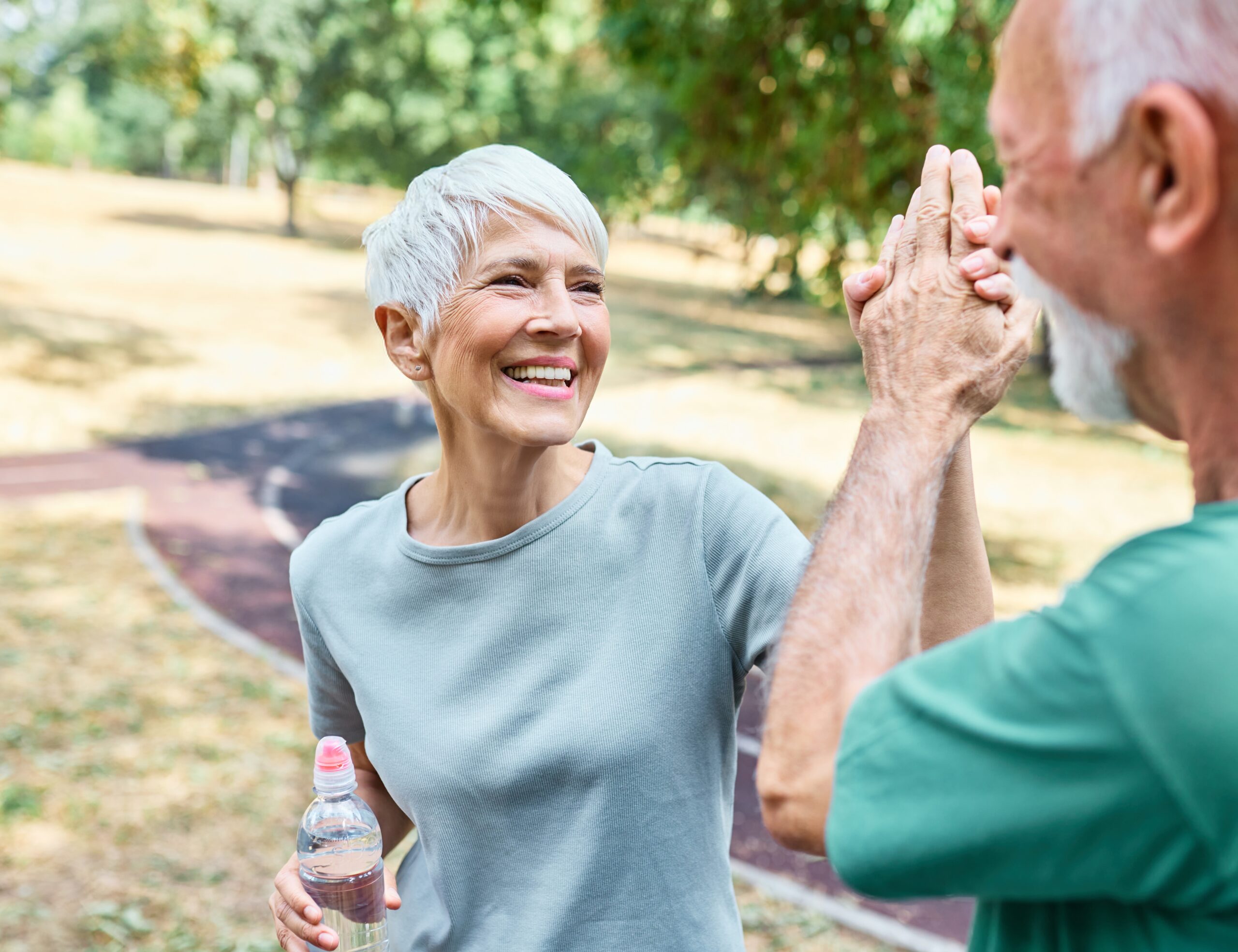 Ältere Frau mit grauem T-Shirt hält eine Wasserflasche und gibt einem älteren Mann im Freien fröhlich einen High-Five.